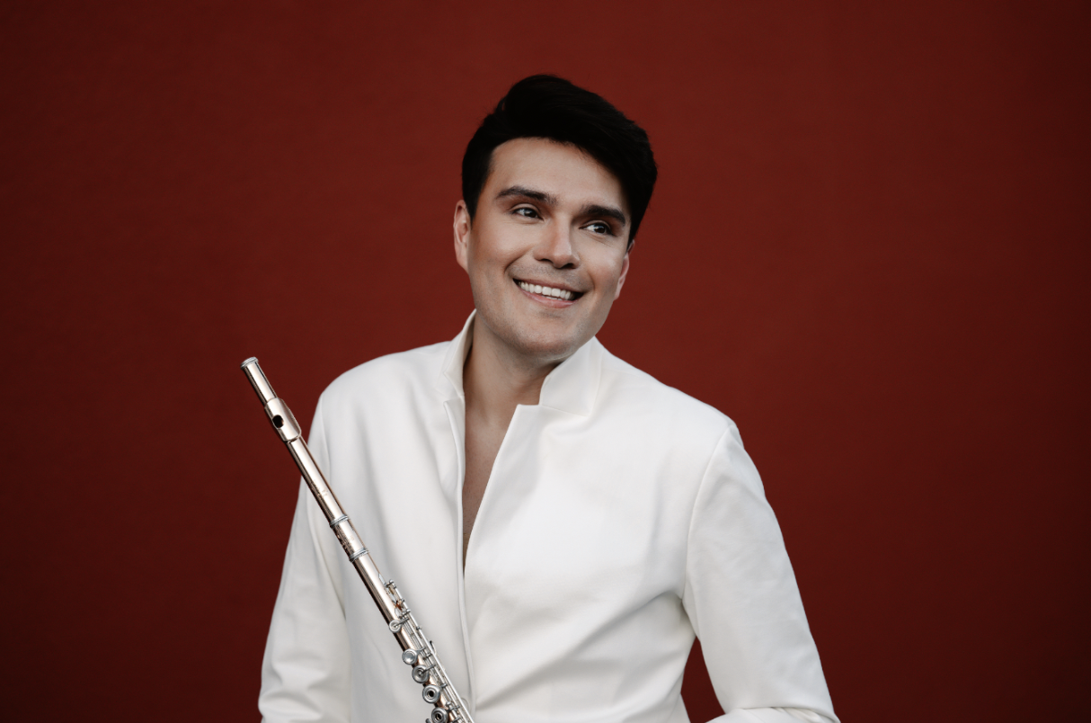 Rayo Furata, holding a flute, smiling and wearing a white dressy shirt with a red background behind him.